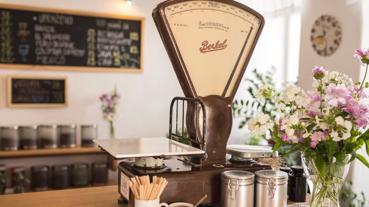A vintage Berkel coffee scale on the counter at Pražírna kávy IDEAL, surrounded by bean jars, fresh flowers, and a handwritten menu board